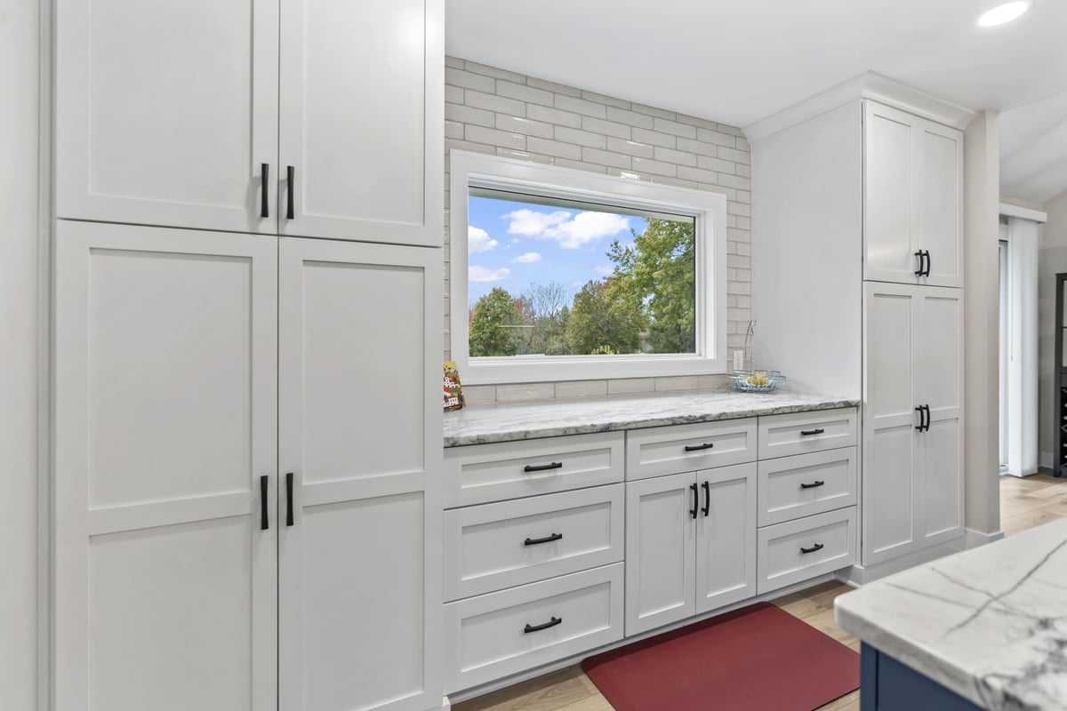 Generous kitchen storage in Stilwell, KS with tall white cabinets, quartz countertops, subway tile backsplash, and large window.